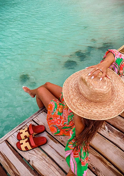 Woman sitting on a wooden pontoon, her feet in the turquoise water, wearing a straw hat and a colorful dress in the Caribbean