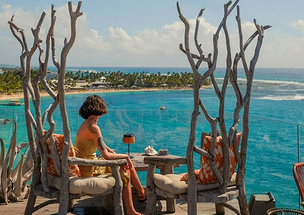 Woman sitting on a wooden pontoon, her feet in the turquoise water, wearing a straw hat and a colorful dress in the Caribbean