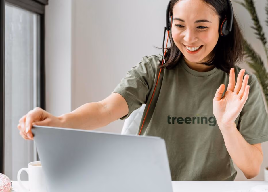 Smiling woman wearing headset waves while looking at laptop during a video call.