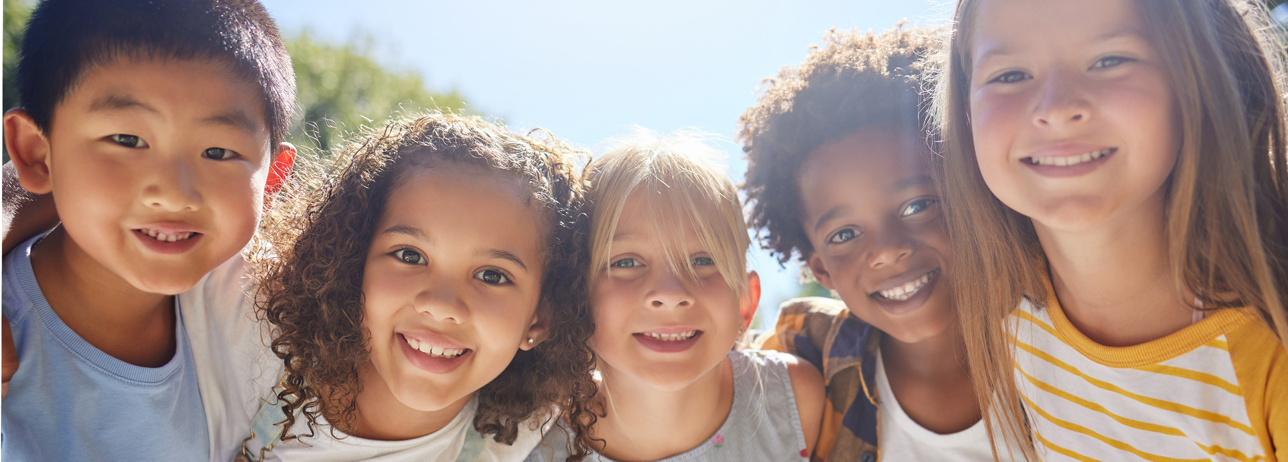 Smiling diverse group of five young children standing outdoors close together under sunlight.