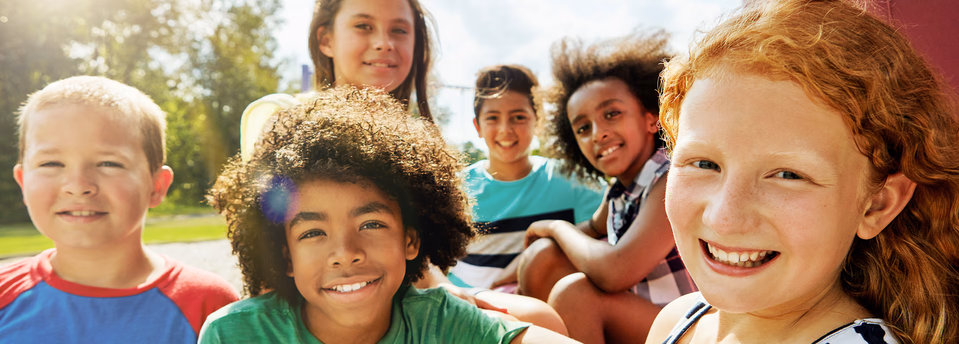 Group of five diverse smiling children outdoors, close together with bright sunlight behind them.