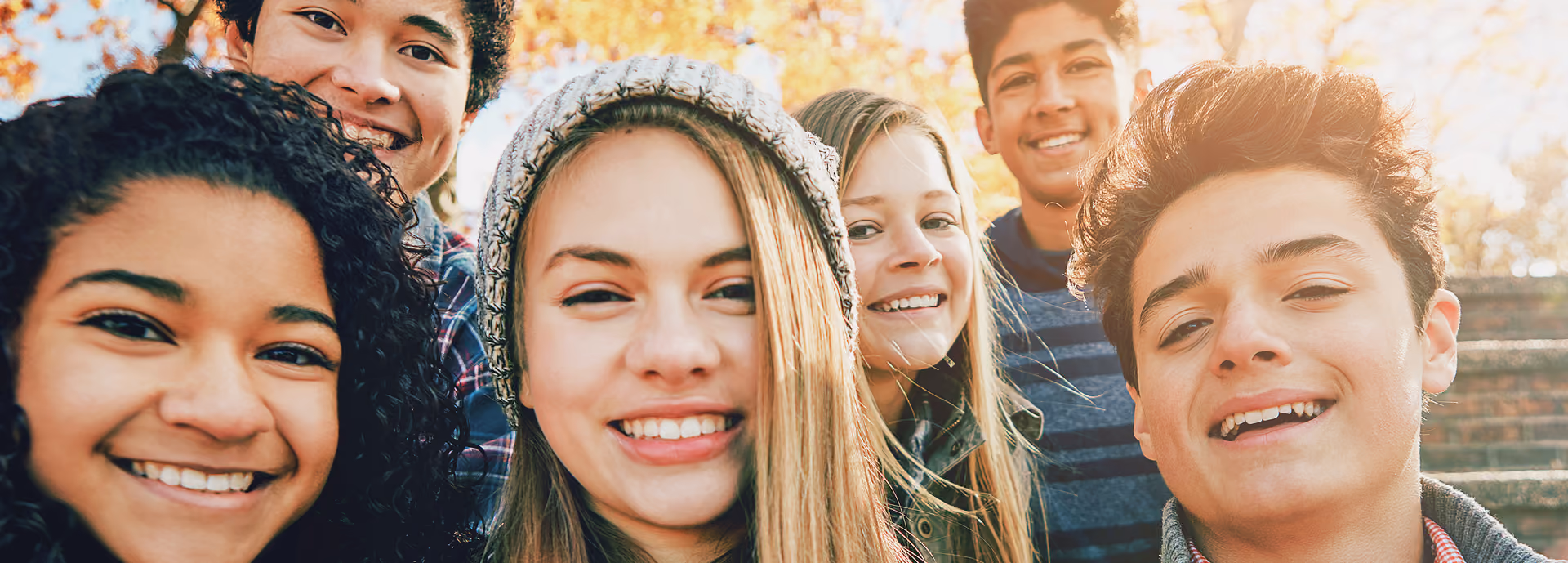 Group of six diverse teens smiling outdoors on a sunny autumn day.