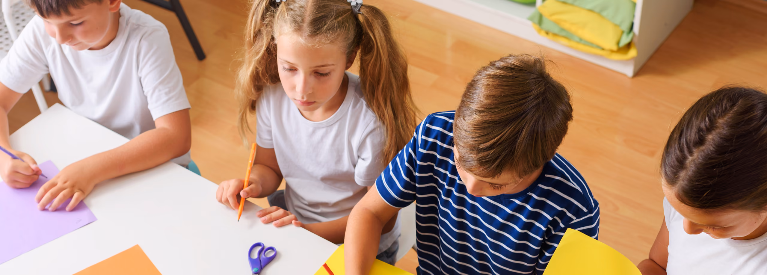 Four children seated at a table drawing and coloring on colored paper with pencils and scissors.