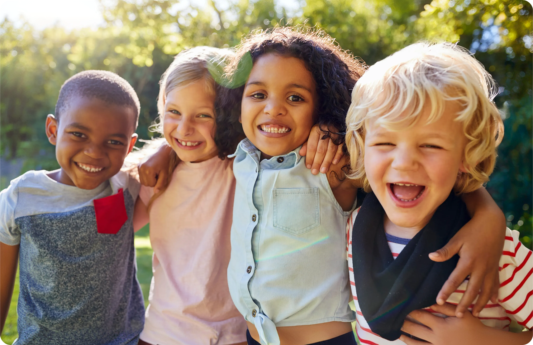 Four diverse children smiling and hugging outdoors with sunlit greenery in the background.