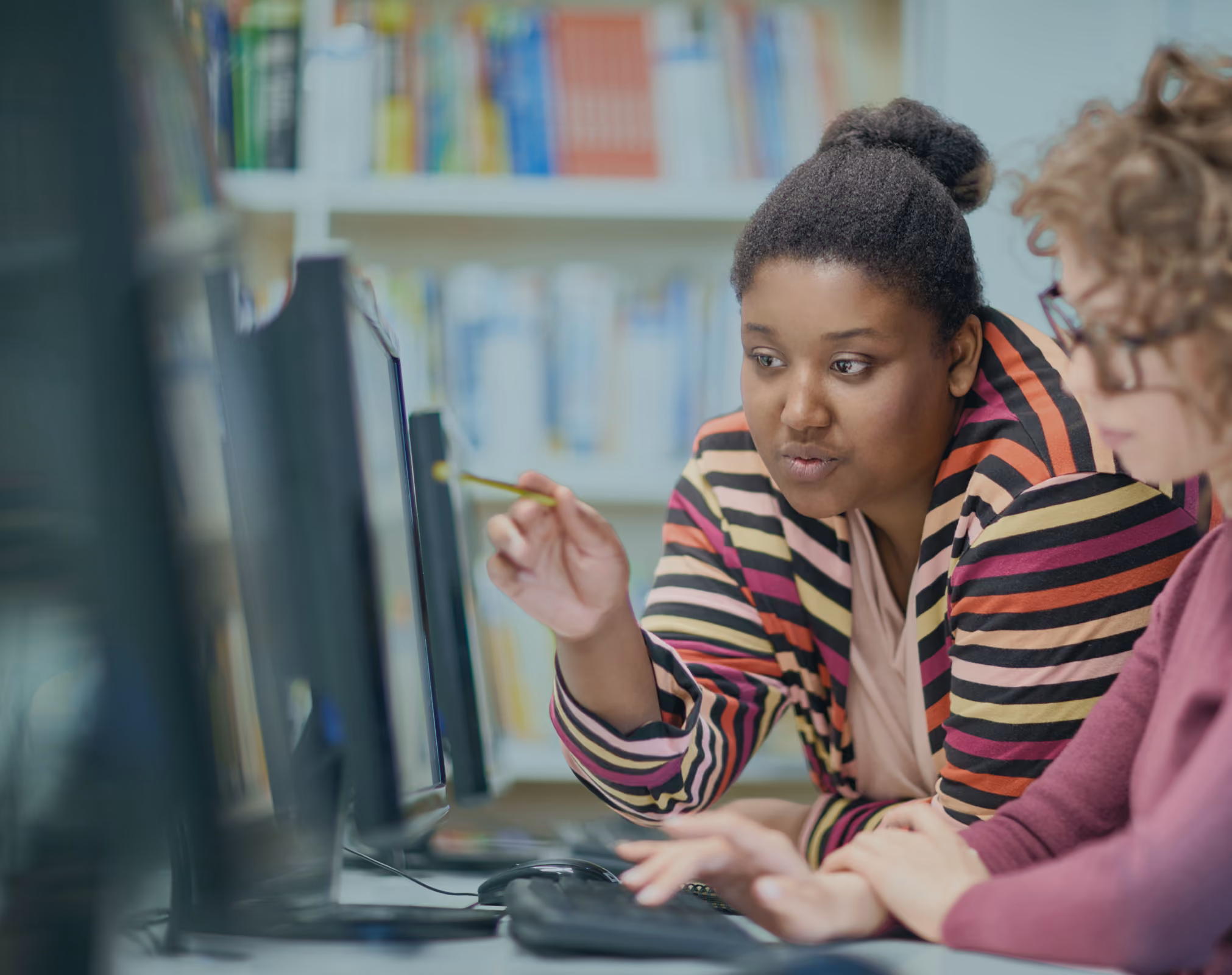 Woman in striped sweater pointing at computer screen while explaining to another woman typing.