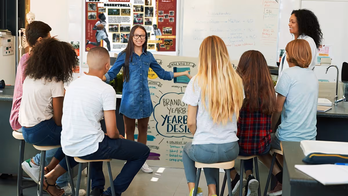 Student presenting yearbook design ideas to classmates and teacher in a classroom.