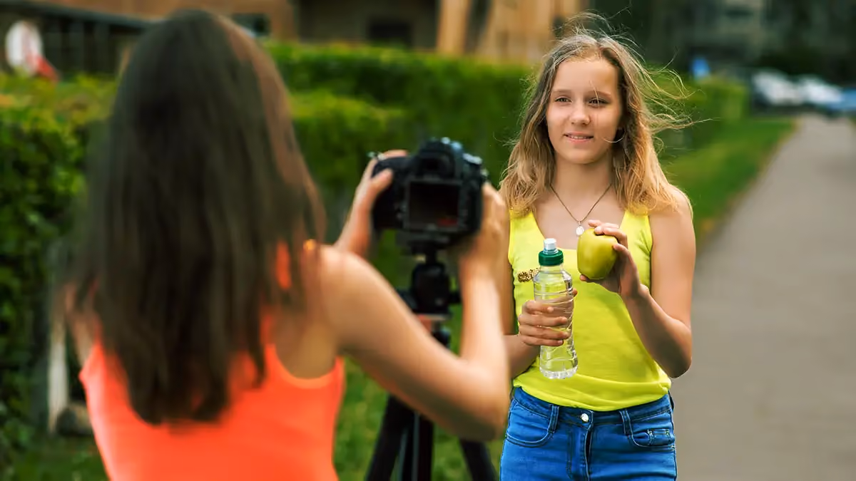 Girl in yellow tank top holding an apple and water bottle, photographed outdoors with another girl.
