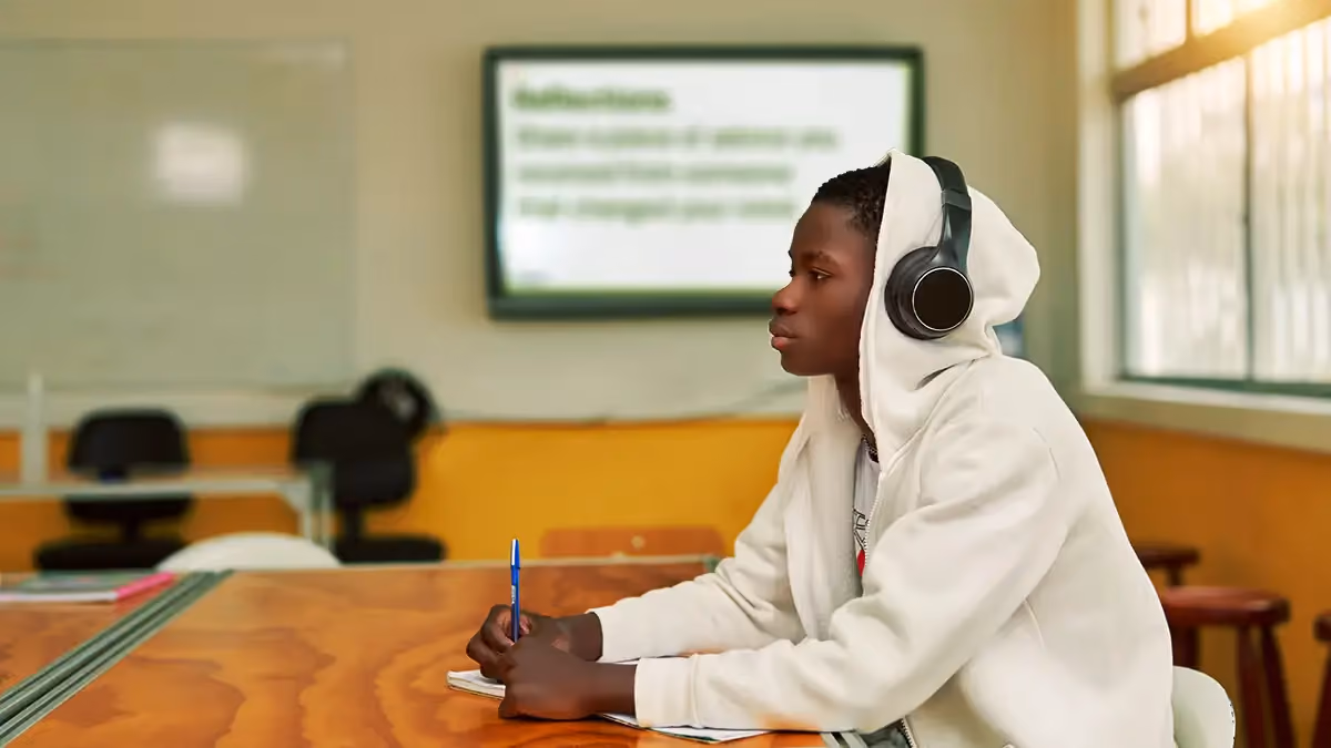 Teen boy wearing headphones and a hoodie sits at a desk in a classroom, writing with a pen.