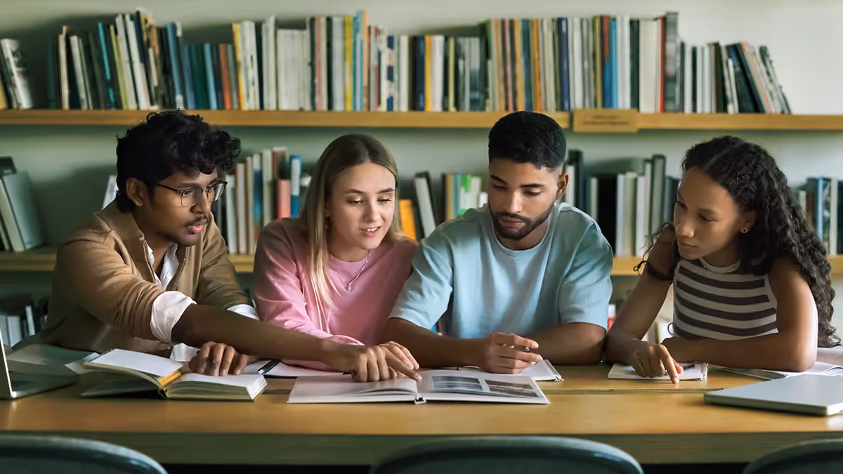 Four diverse students sitting at a table, studying together with books in a library.