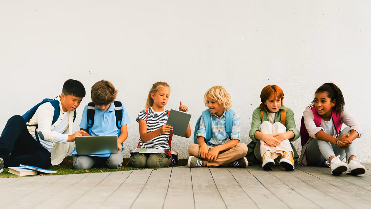 Six children sitting against a white wall, using a laptop and tablets with backpacks on.