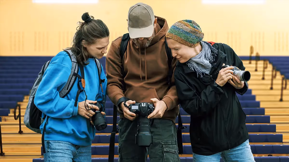 Three photographers reviewing photos on a digital camera in an indoor stadium with blue and yellow bleachers.
