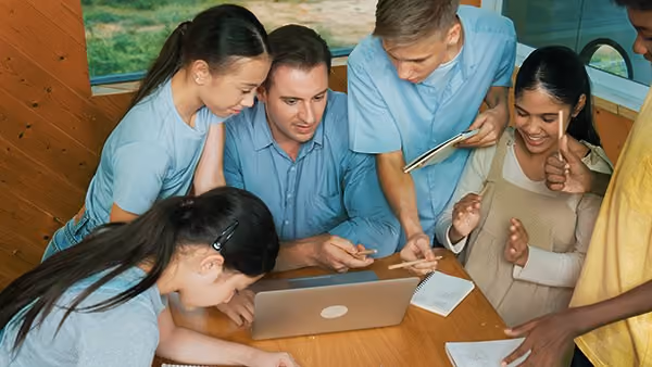 Group of young people gathered around a laptop, engaged in discussion and note-taking at a wooden table.