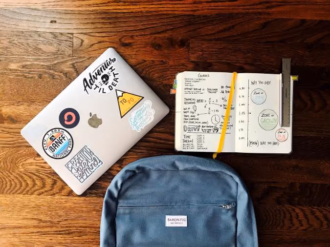 Laptop with stickers, open notebook with notes and pencil, and blue Baron Fig backpack on wooden floor.