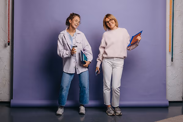 Two young women standing in front of a purple backdrop holding notebooks and a coffee cup, smiling.