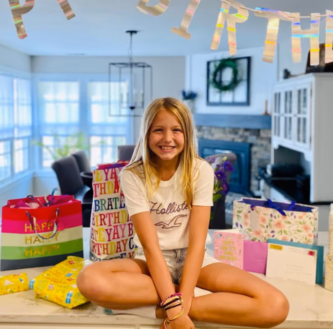 Smiling girl sitting cross-legged on a kitchen counter with birthday gifts and decorations around her.