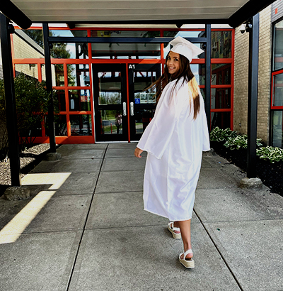 Photo of a senior in her cap and gown walking towards the school building, looking over her shoulder