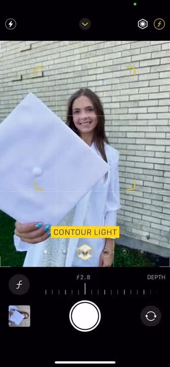 Young woman in white holds a white graduation cap toward the camera, brick wall background.