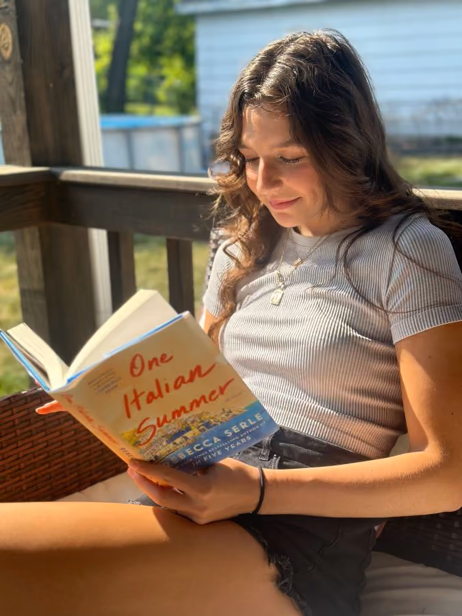 Young woman sitting outdoors reading the book 'One Italian Summer' by Rebecca Serle in sunlight.