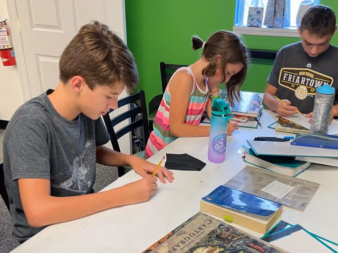 Three students sitting at a table writing with pencils, surrounded by books and water bottles.
