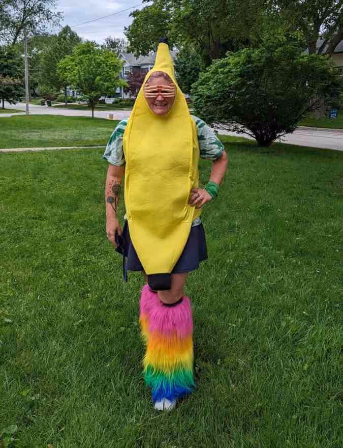 Person in banana costume with rainbow-colored furry leg warmers standing on grass outdoors.