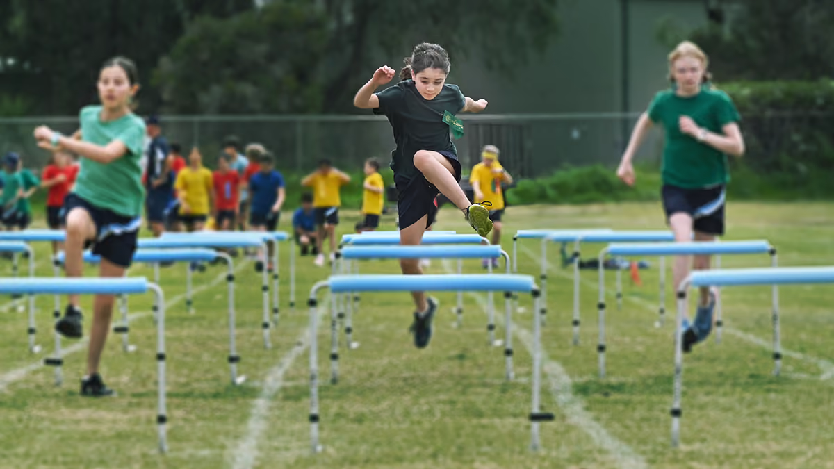Children in sportswear jumping over hurdles on a grassy field during a race.