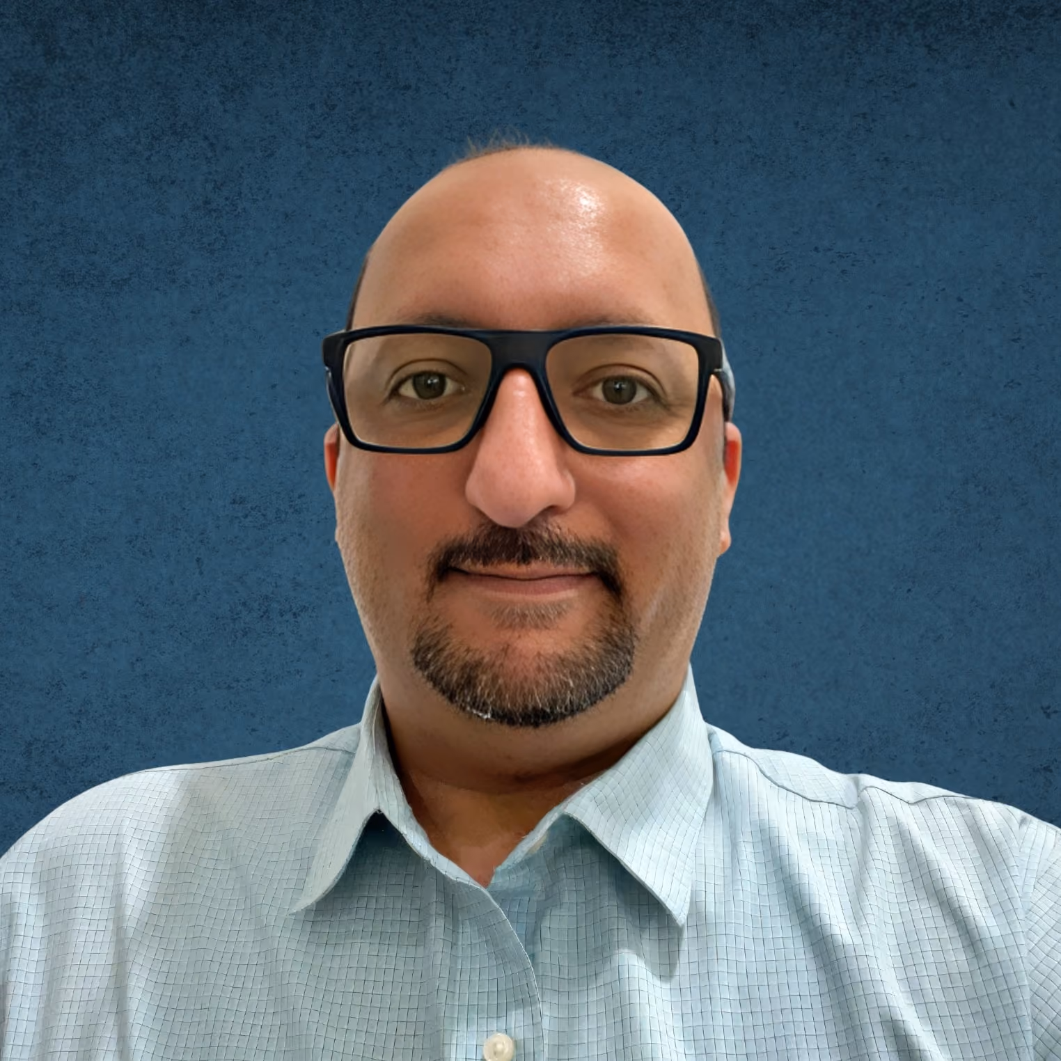 Smiling bald man with glasses and a goatee wearing a light blue checkered shirt against a blue background.