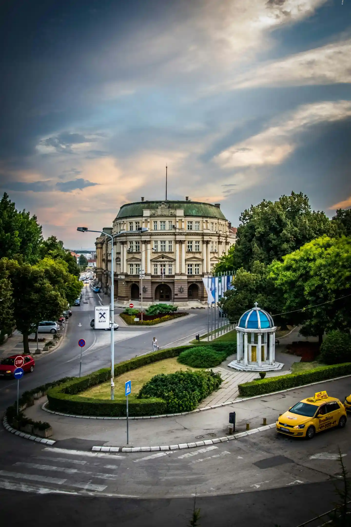Ulica sa zelenilom i taksijem u prvom planu, istorijska zgrada i plavo nebo sa oblacima u pozadini.