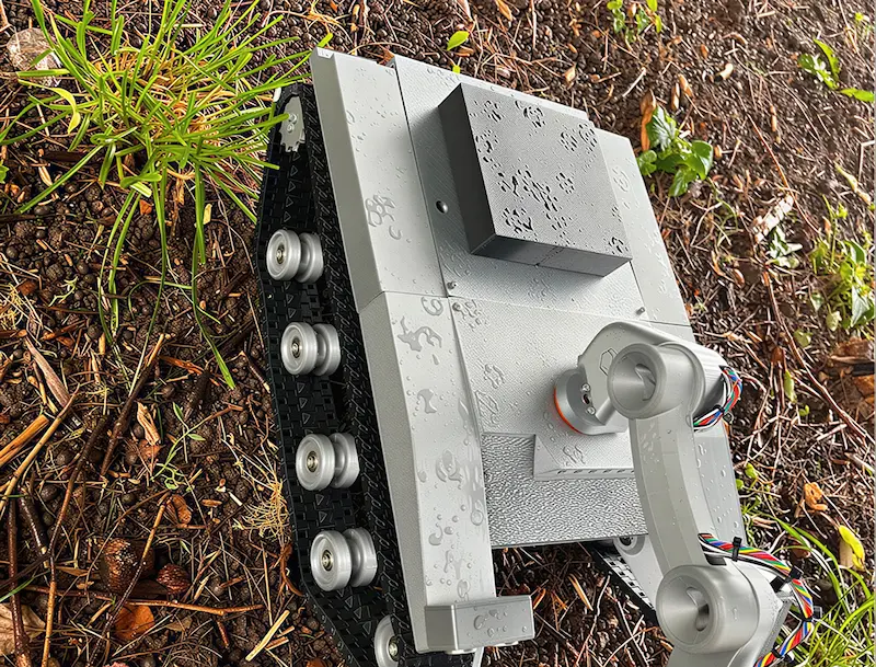 Sivi robotski droid na guseničaru sa krakom, na vlažnoj zemlji sa lišćem i travom.