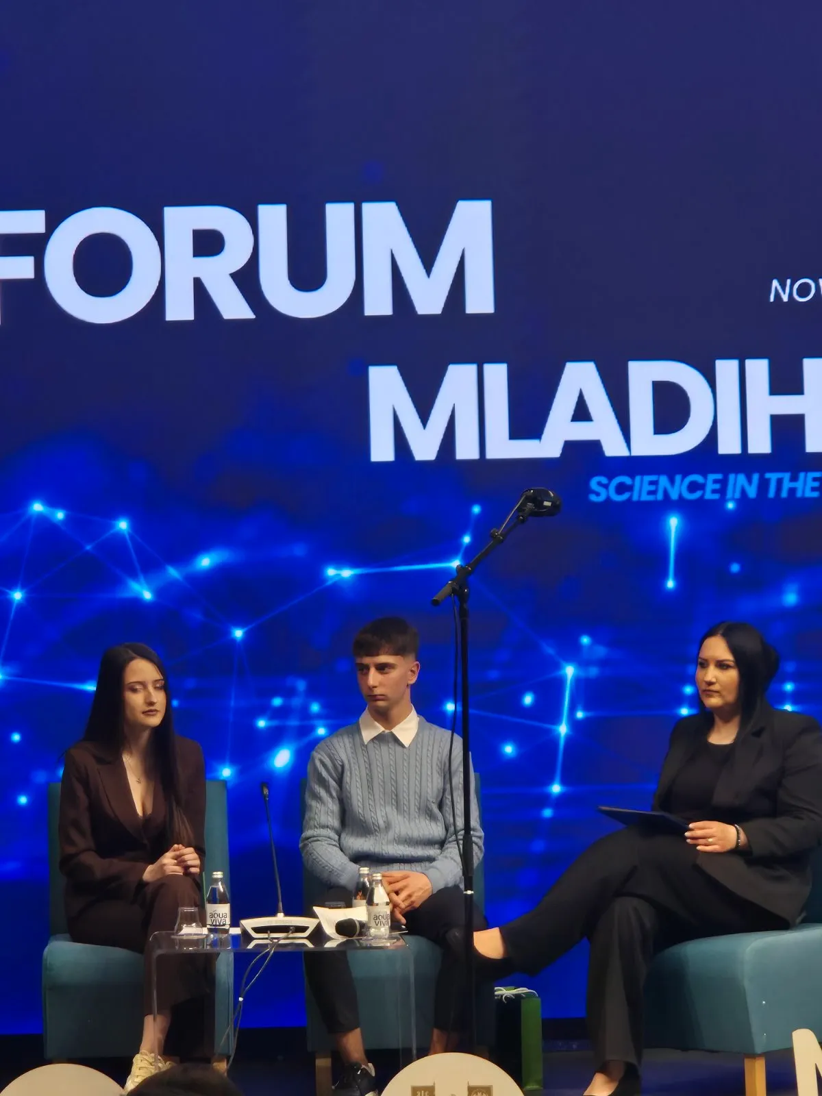 Tri osobe sede na pozornici na forumu o mladima sa plavim digitalnim ekranom u pozadini.