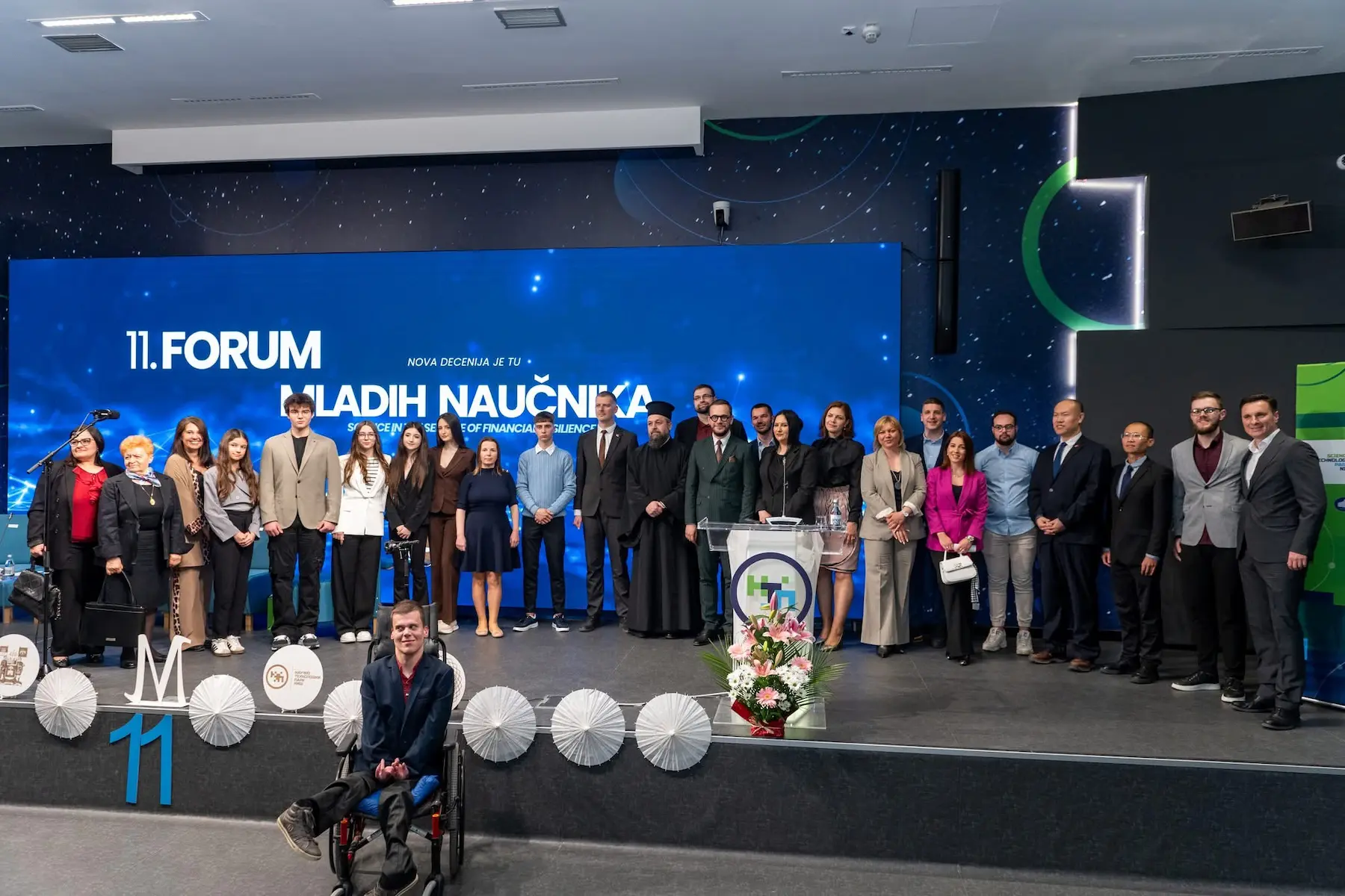 Grupa ljudi pozira na sceni foruma mladih naučnika sa plavim ekranom u pozadini.