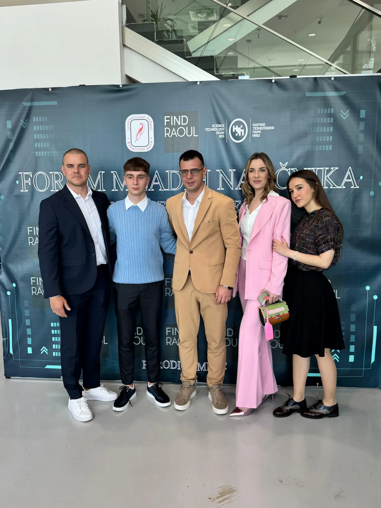 Grupa od pet ljudi stoji ispred banera Foruma mladih naučnika u poslovnom prostoru.