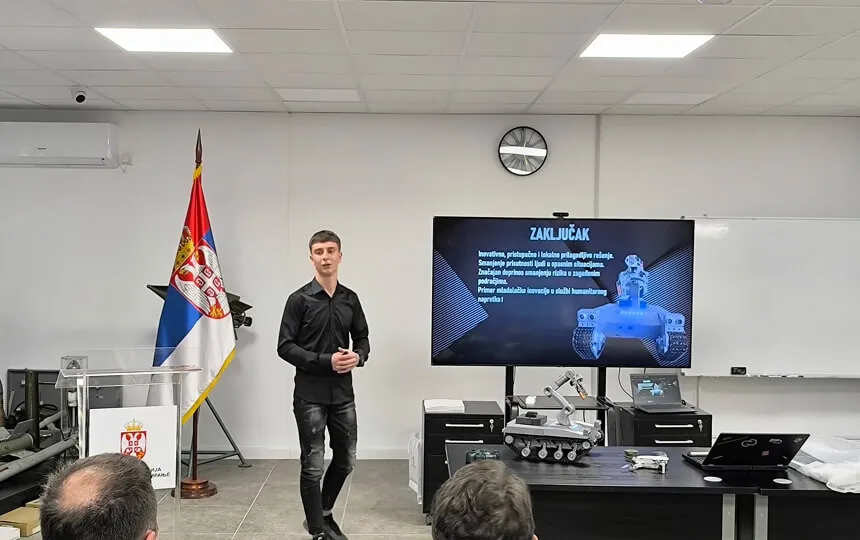 Mladić drži prezentaciju sa modelom robota i zastavom Srbije u pozadini.