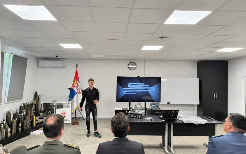 Muškarac drži prezentaciju pred tri muškarca u vojnim uniformama sa srpskom zastavom i izložbenim predmetima.