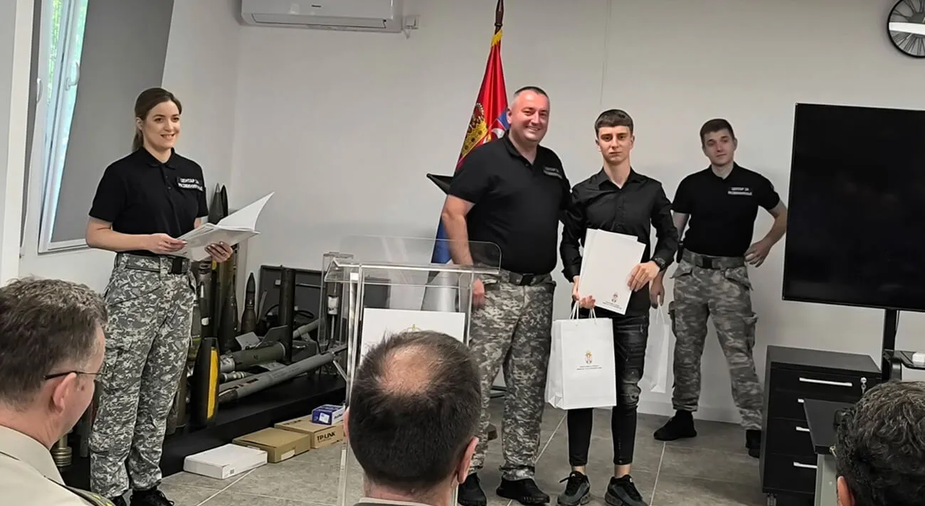 Četvoro ljudi u uniformi u kancelariji, dvoje drže dokumenta i kese pred publiku.