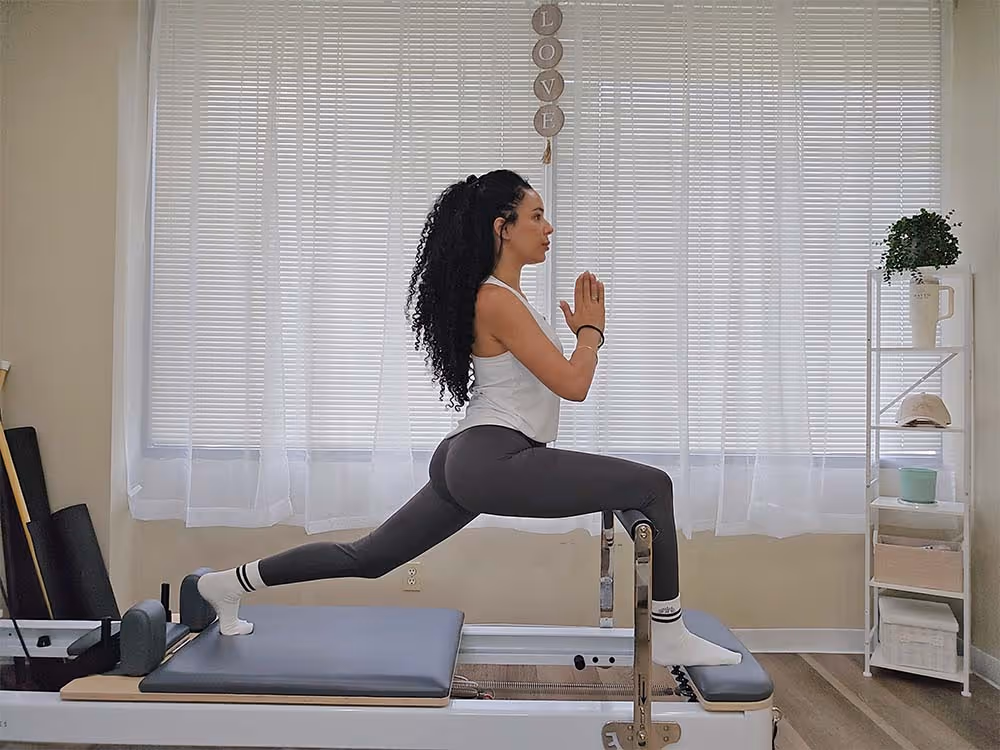 Woman with curly hair in athletic wear doing a lunge stretch on a Pilates reformer machine in a bright room with white curtains and a shelf with plants and decor.