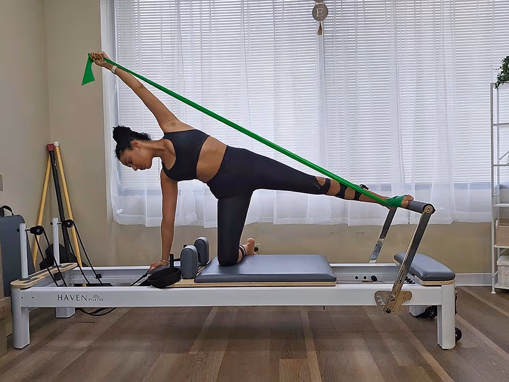 Woman in black workout clothes using a green resistance band on a Pilates reformer machine in a bright room.