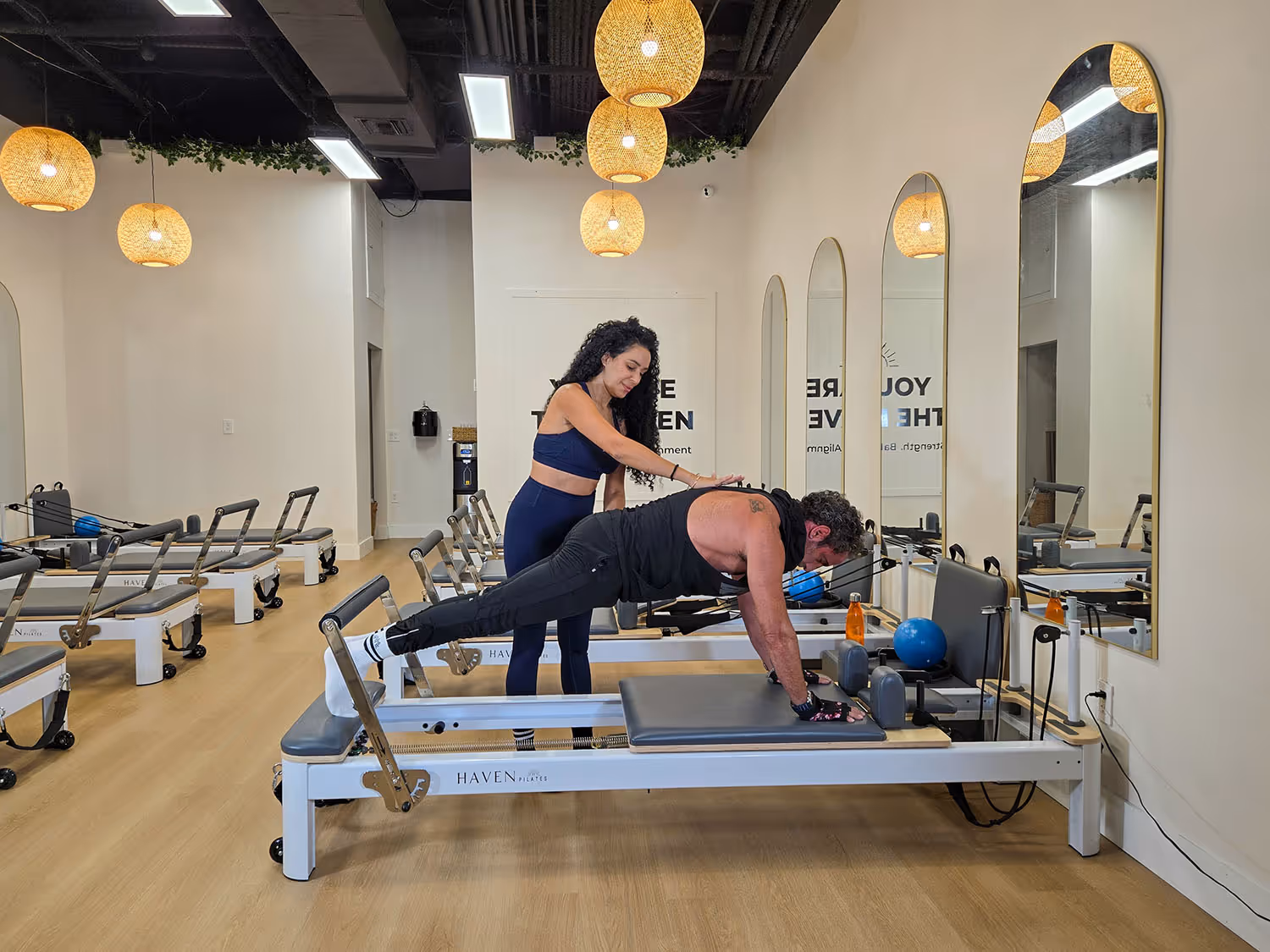 Female instructor assists a man doing a plank exercise on a Pilates reformer machine in a bright studio.