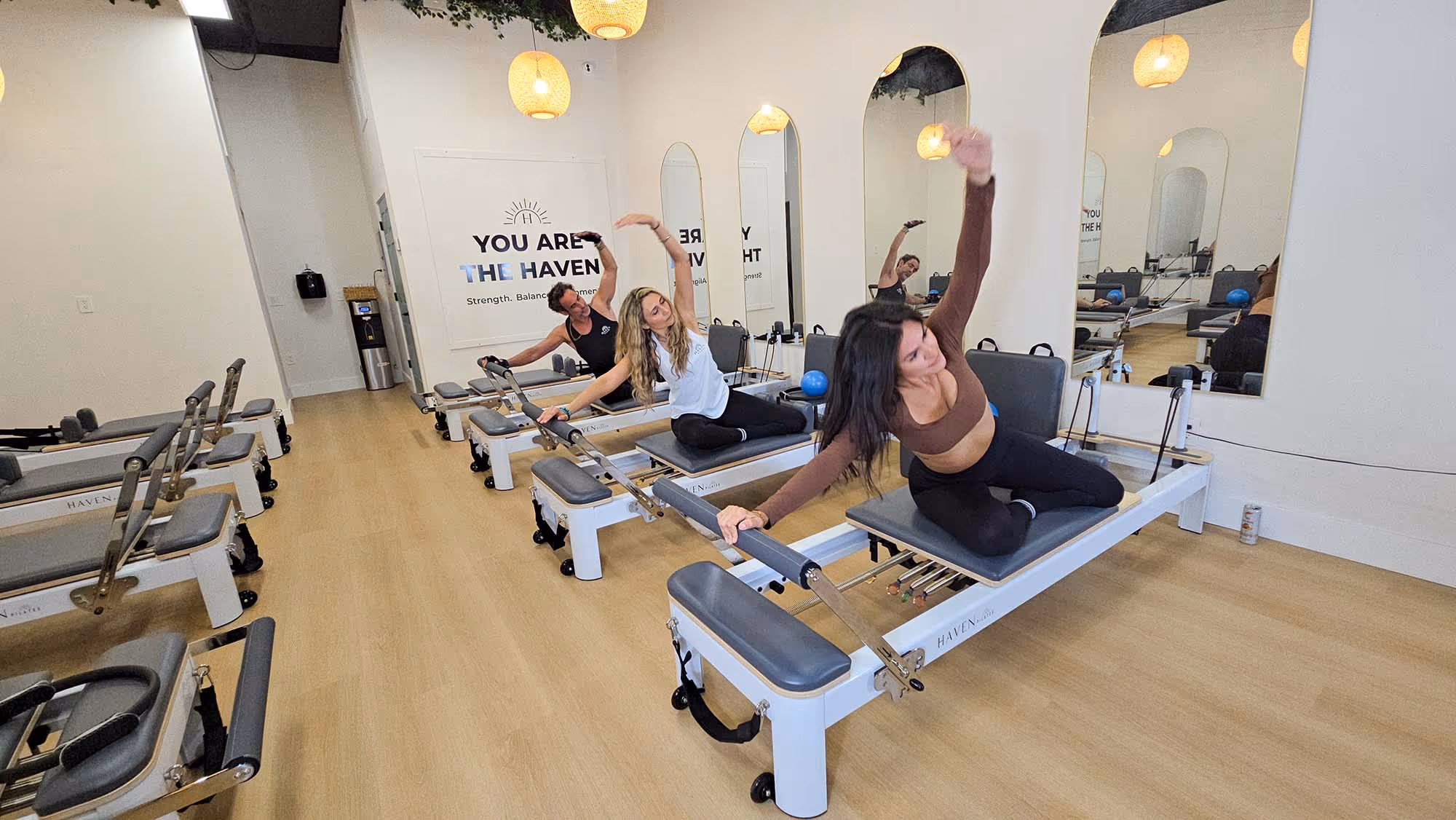 Three people exercising on Pilates reformer machines in a bright studio with wooden floors and arched mirrors.