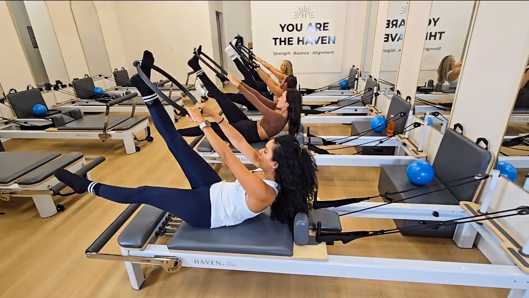 Four women exercising on reformer machines in a Pilates studio with a sign reading 'You Are The Haven'