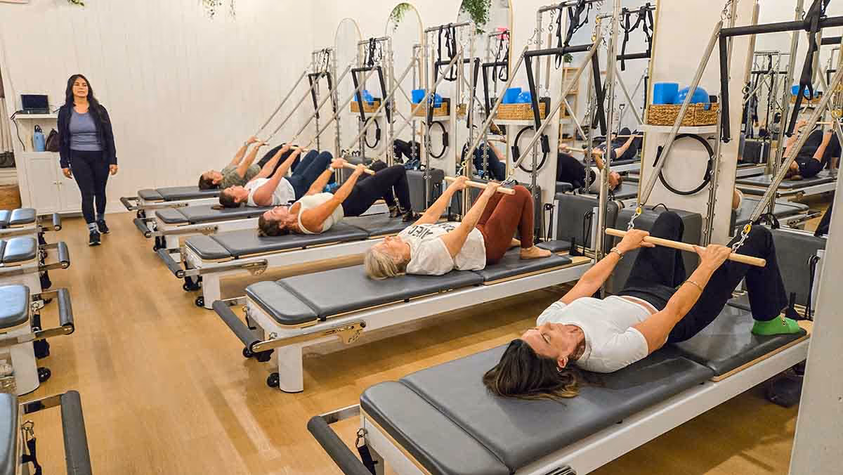 Women exercising on reformer Pilates machines in a studio while instructor watches.