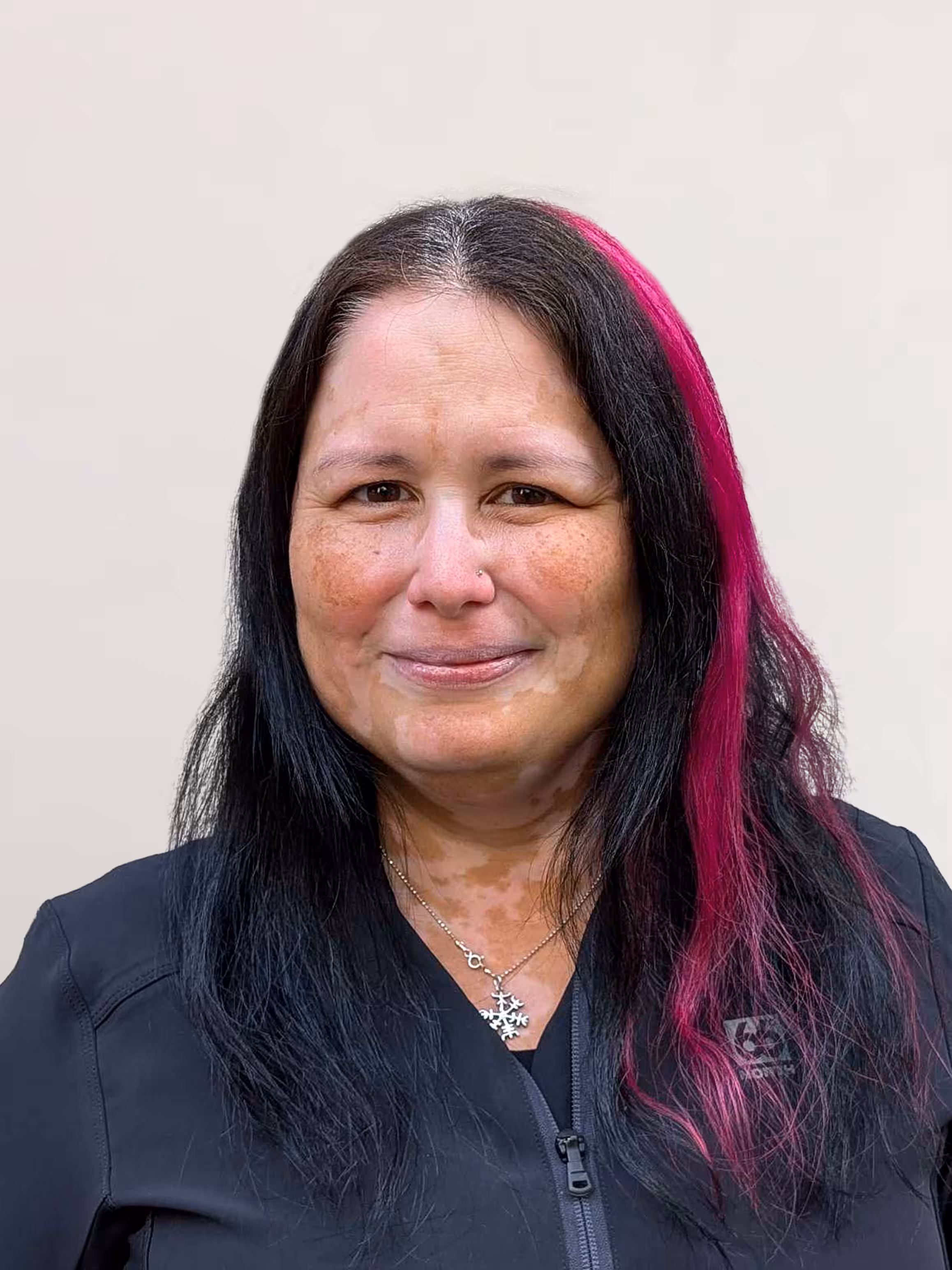 Smiling woman with long black hair featuring a bright pink streak, wearing a black zip-up top and a snowflake necklace.