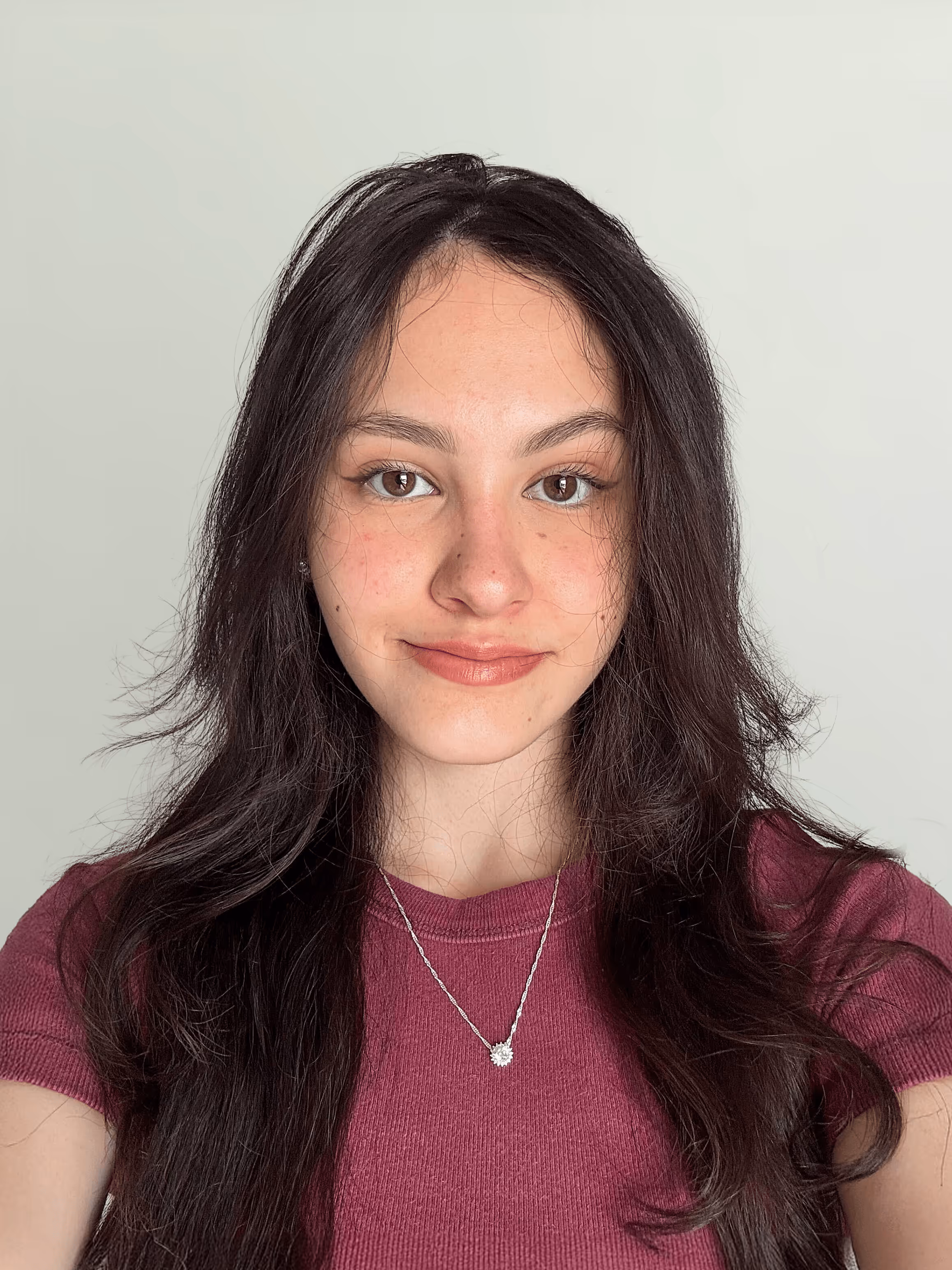 Young woman with long dark hair wearing a purple shirt and silver pendant necklace against a plain light background.
