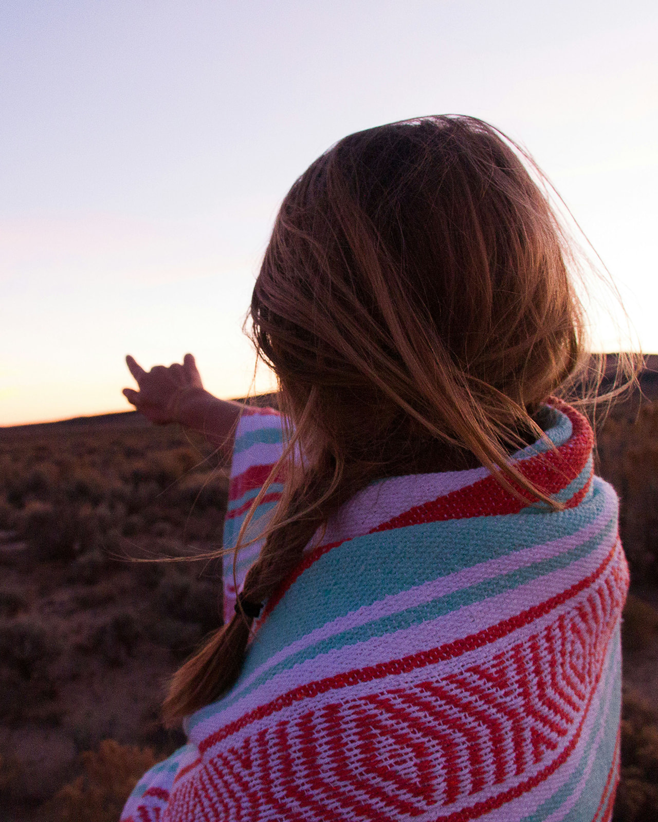 Woman with braided hair wrapped in a colorful blanket pointing towards a sunset in a desert landscape.