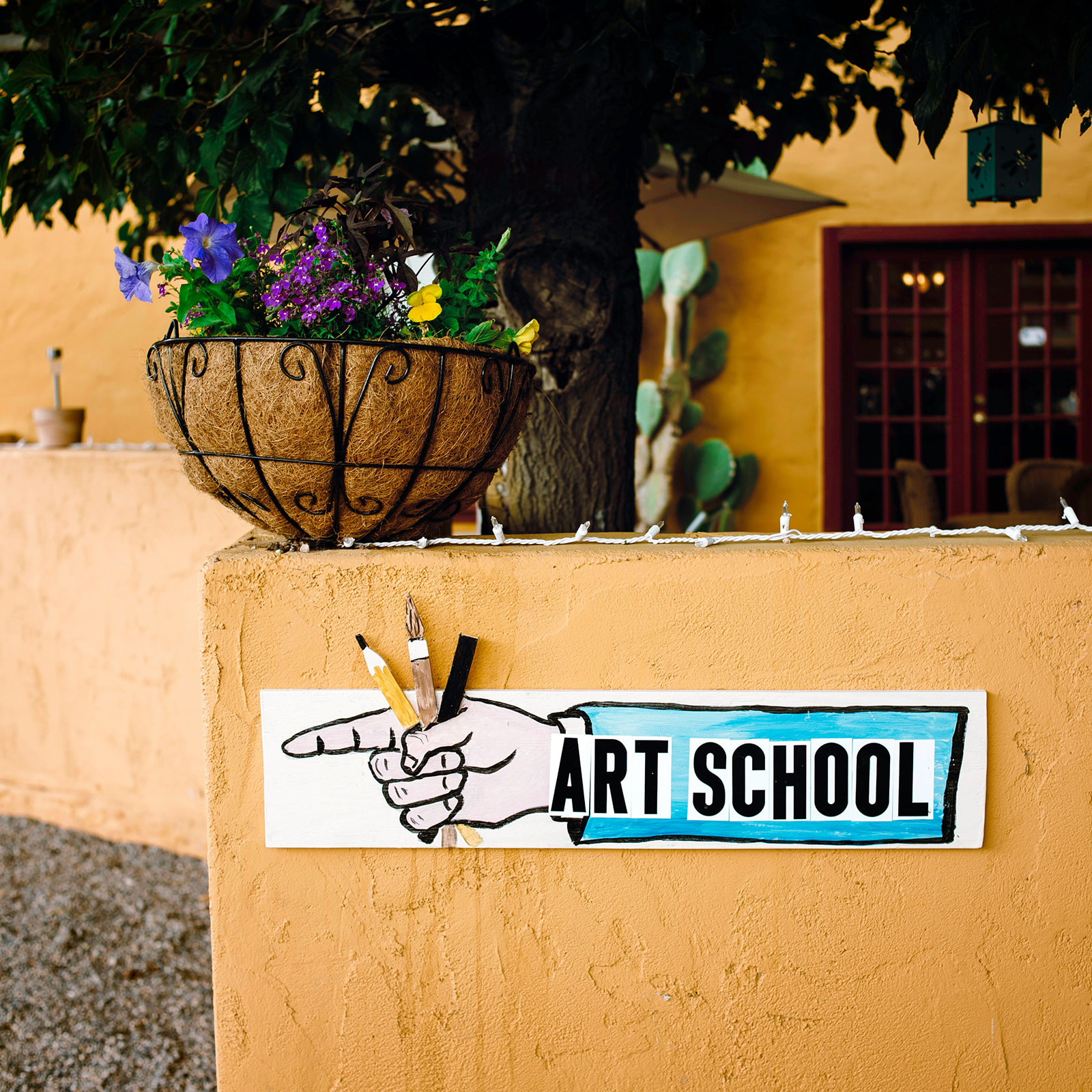 Hand-painted sign with a pointing hand holding art tools directing to an art school, with a hanging basket of flowers above on a textured yellow wall.