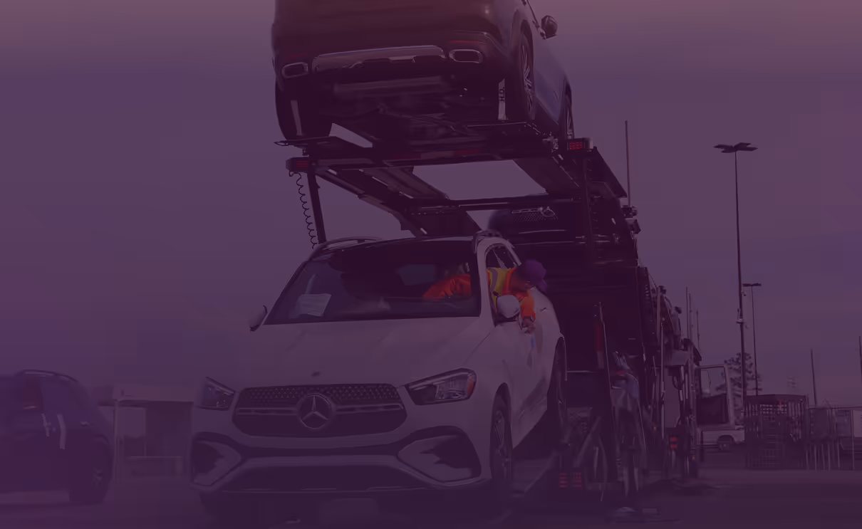 Worker in orange and yellow safety vest leaning out of a white Mercedes SUV loaded on a car carrier truck in a parking lot.