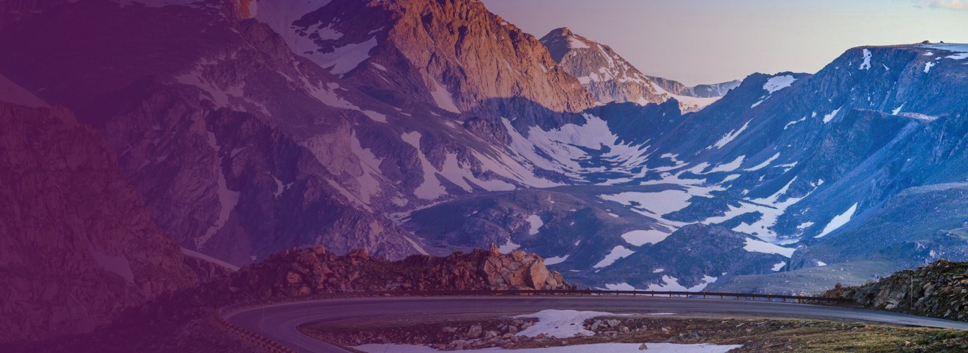 Twisting mountain road with surrounding rocky peaks and patches of snow at sunset.
