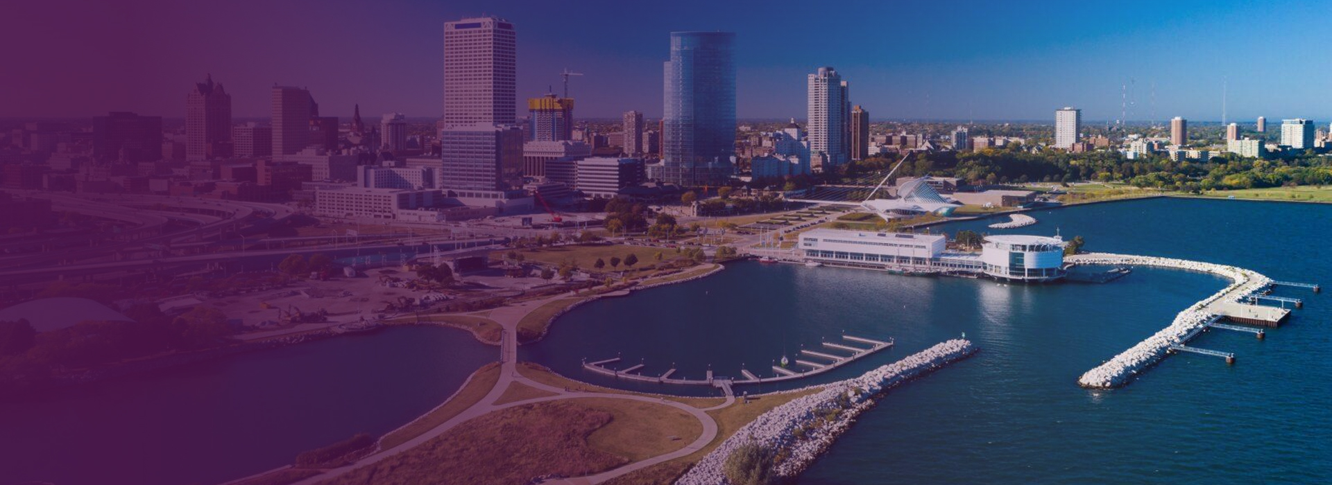 Aerial view of Milwaukee skyline with lakeside harbor, walking paths, and the Milwaukee Art Museum under clear blue sky.