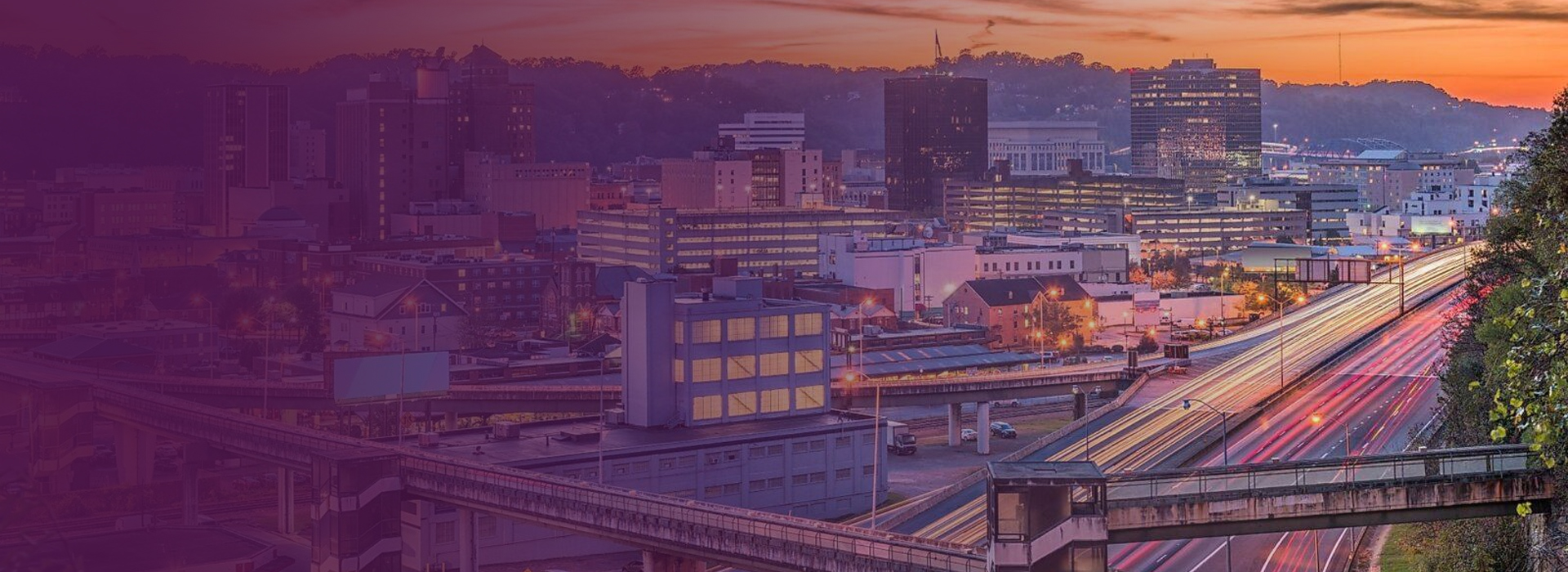 Cityscape of Charleston, West Virginia at sunset with illuminated buildings and light trails on a busy highway.