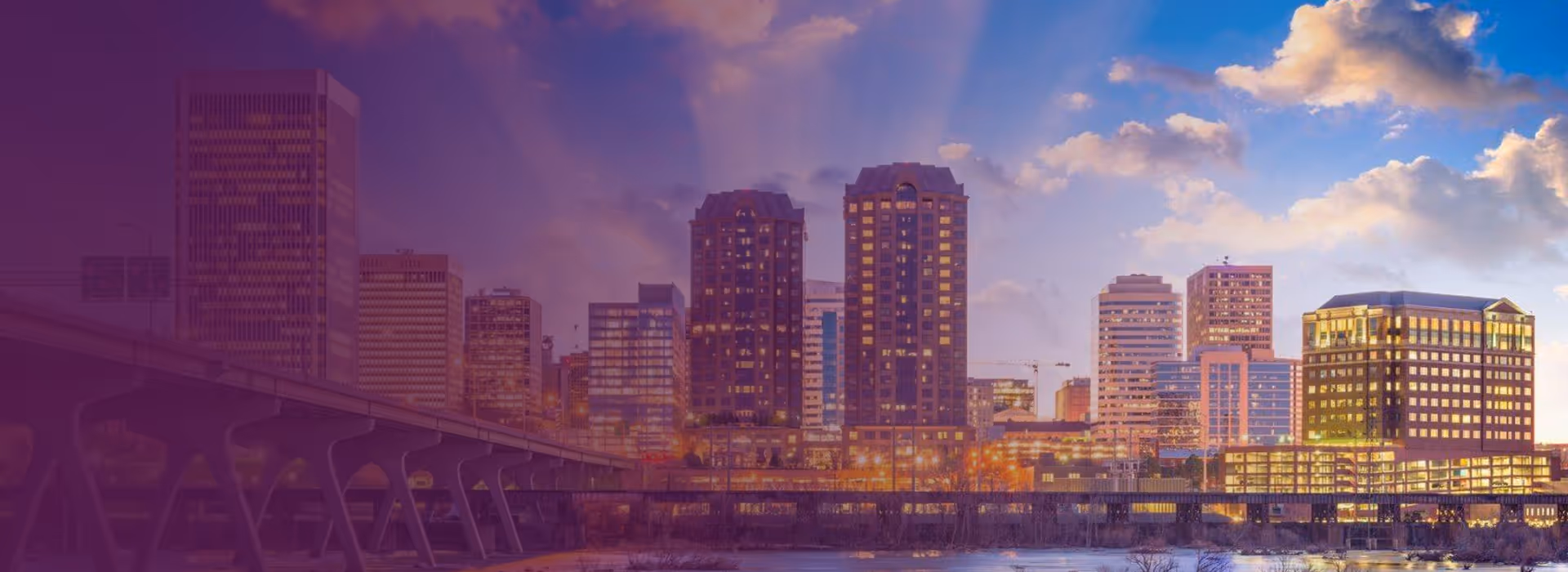 Sunset view of a city skyline with illuminated buildings and a bridge over a body of water.