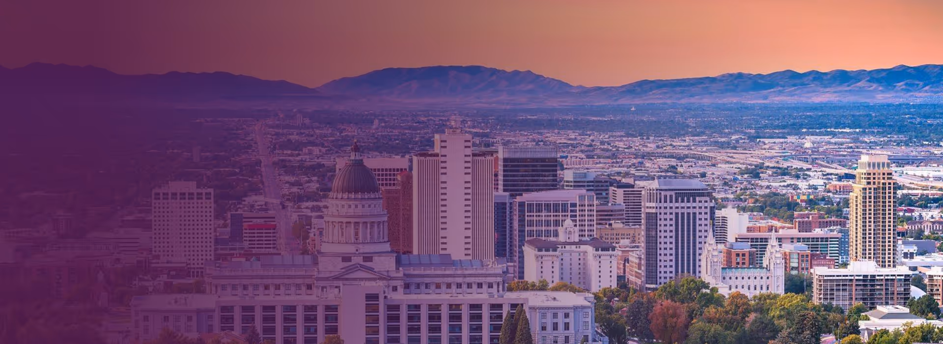 City skyline with Utah State Capitol building in foreground and mountains in the background at sunset.